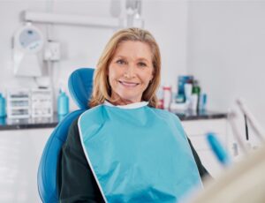 Old beautiful lady smiling while seated on dental chair wearing a blue bib for dental implant. Confident elderly patient waiting for whitening treatment in clinic, dentures and dental prosthesis.