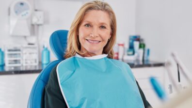 Old beautiful lady smiling while seated on dental chair wearing a blue bib for dental implant. Confident elderly patient waiting for whitening treatment in clinic, dentures and dental prosthesis.